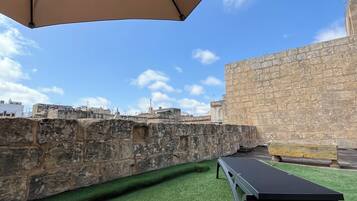 An outdoor area with a stone wall, green artificial turf, and two lounge chairs under a large umbrella.