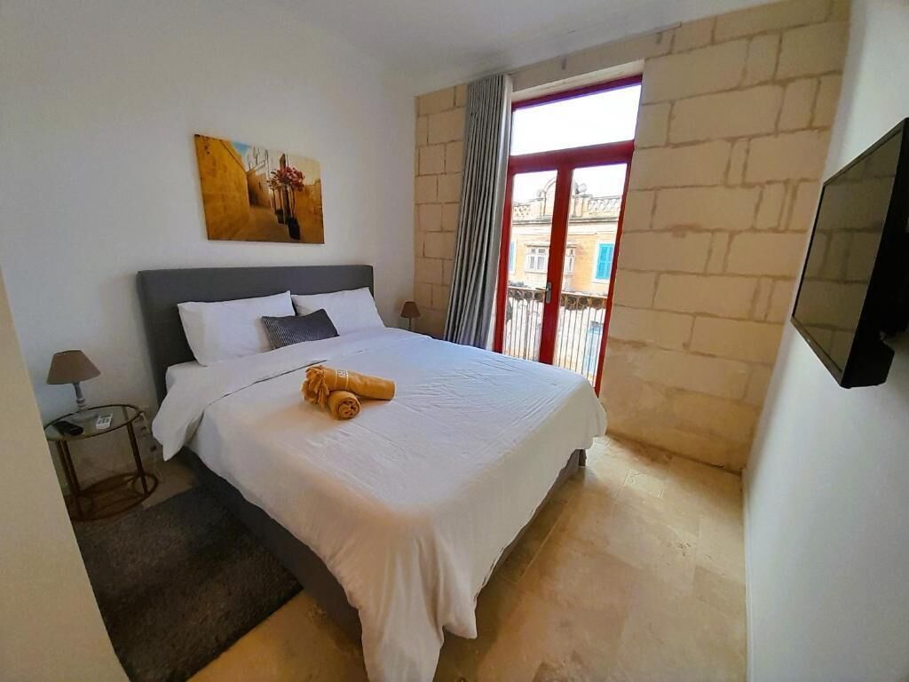 A bedroom with a bed, a nightstand, a lamp, a painting, a window with a view, and a TV mounted on the wall.