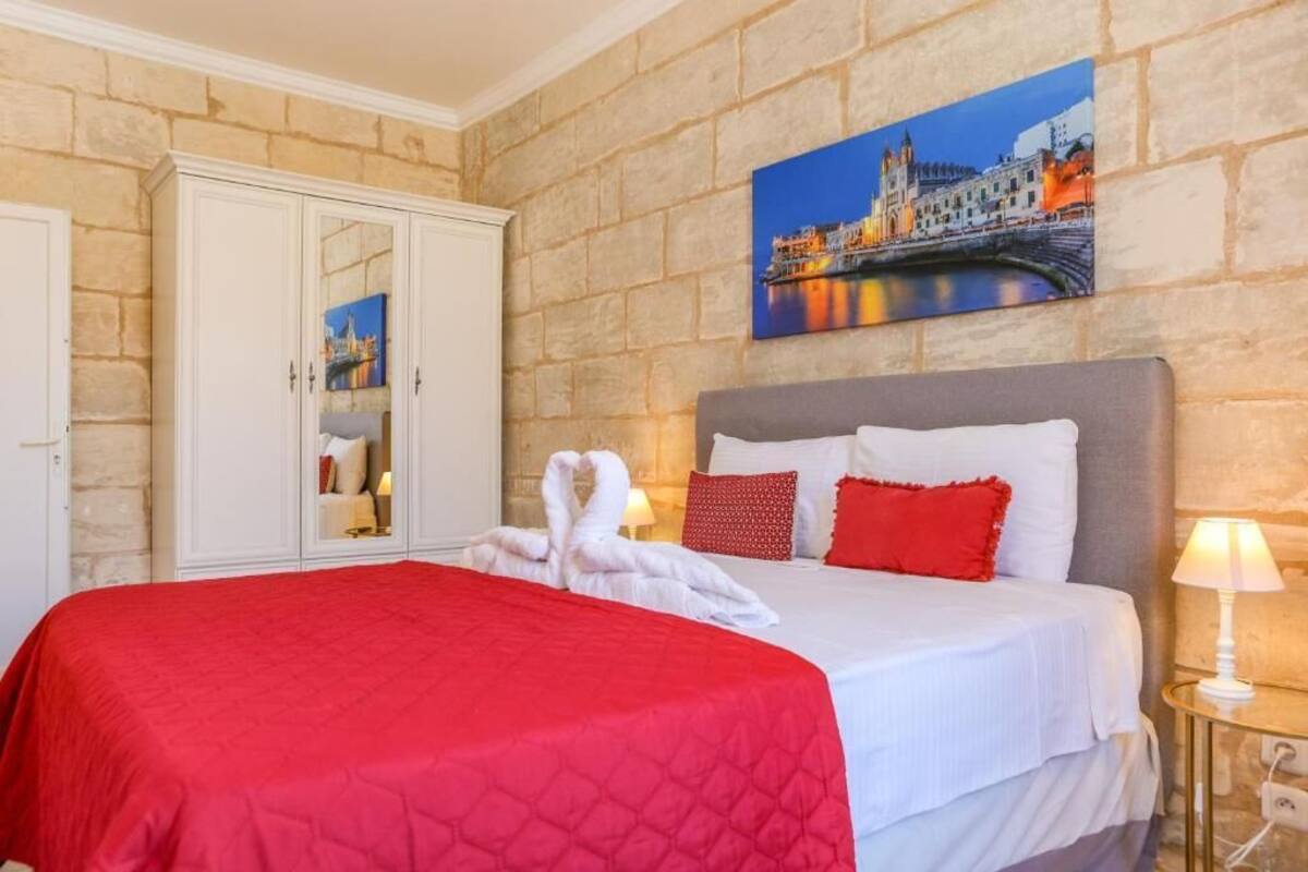 A hotel room with a stone wall, a bed with red bedding, a painting of a cityscape, and a wardrobe.