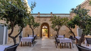 An outdoor dining area with tables and chairs, a stone building with an arched entrance, and trees. - Palazzo Pisani Malta Boutique & Spa (Zabbar)