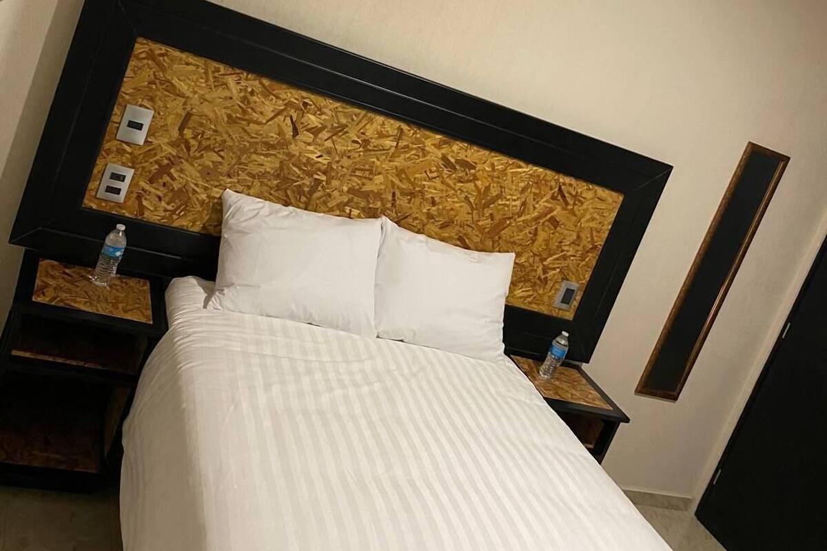 Habitación de hotel con cama, cabecera de diseño texturizado, dos mesitas de noche con botellas de agua y un espejo en la pared.