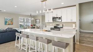 A modern kitchen with white cabinets, a central island with bar stools, stainless steel appliances, and a dining area with a sofa and table.