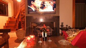 Un salon chaleureux avec une cheminée, un téléviseur fixé au-dessus et une table avec des verres à vin et une bouteille.