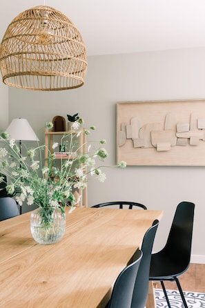 Ein Essbereich mit Holztisch, schwarzen Stühlen, einer Weiden-Pendelleuchte, einer Blumenvase, einer Lampe und einem Holzbilde an der Wand.