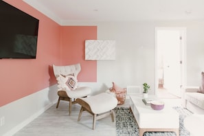 Ein modernes Wohnzimmer mit einer rosafarbenen Akzentwand, einem weißen Sofa, einem weißen Sessel mit Kissen, einem flachbildfernseher, der an der Wand montiert ist, und einem kleinen Topfpflanzen auf einem weißen Tisch.