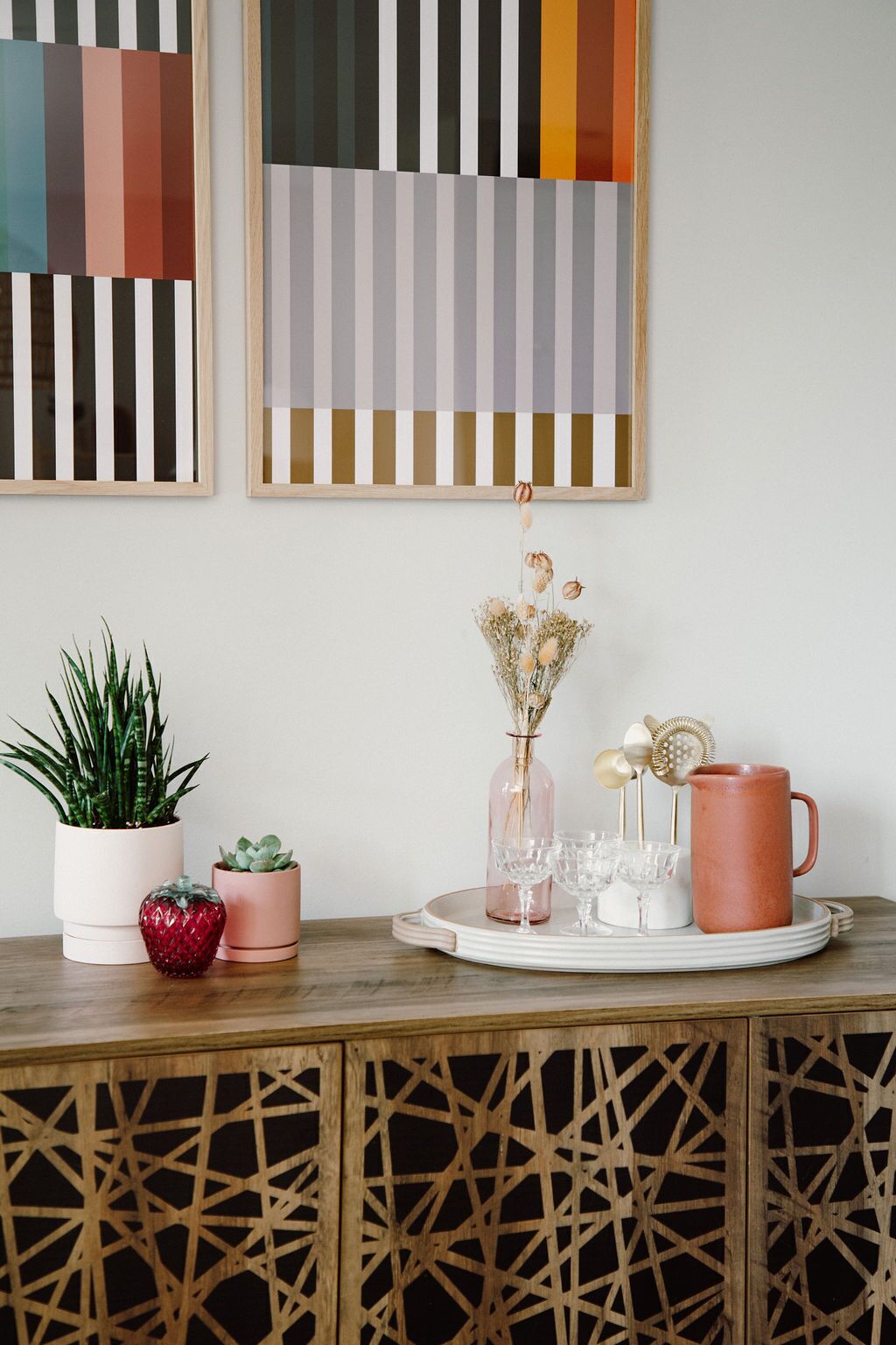 A wooden table with a tray holding a pink vase, a glass pitcher, and a mug. There are two potted plants and a small red object on the table.