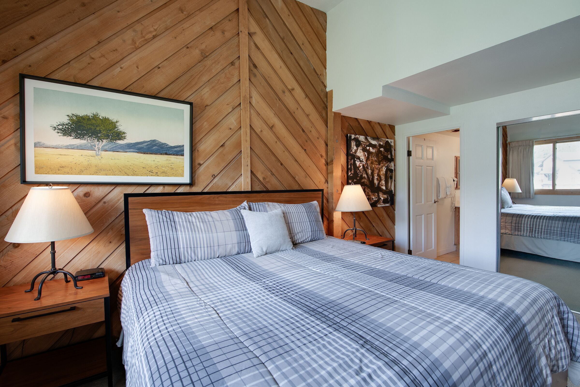 Een slaapkamer met een houten visgraatwand, een bed met gestreept beddengoed, twee bedlampen en een lijst met een landschapsafbeelding.