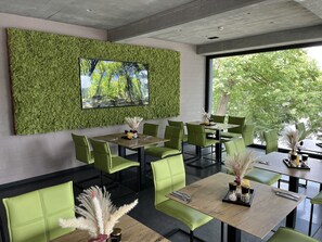 A modern restaurant with green upholstered chairs, a large wall-mounted TV, and a view of trees through the window. - Zweirad Hotel Lenne (Schwerin)