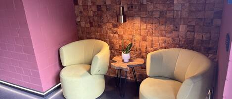 A corner room with two cream-colored armchairs, a small wooden table with a potted plant, and a brick wall with a wooden panel design.