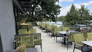 An outdoor patio with tables and chairs, overlooking a body of water and trees. - Zweirad Hotel Lenne (Schwerin)