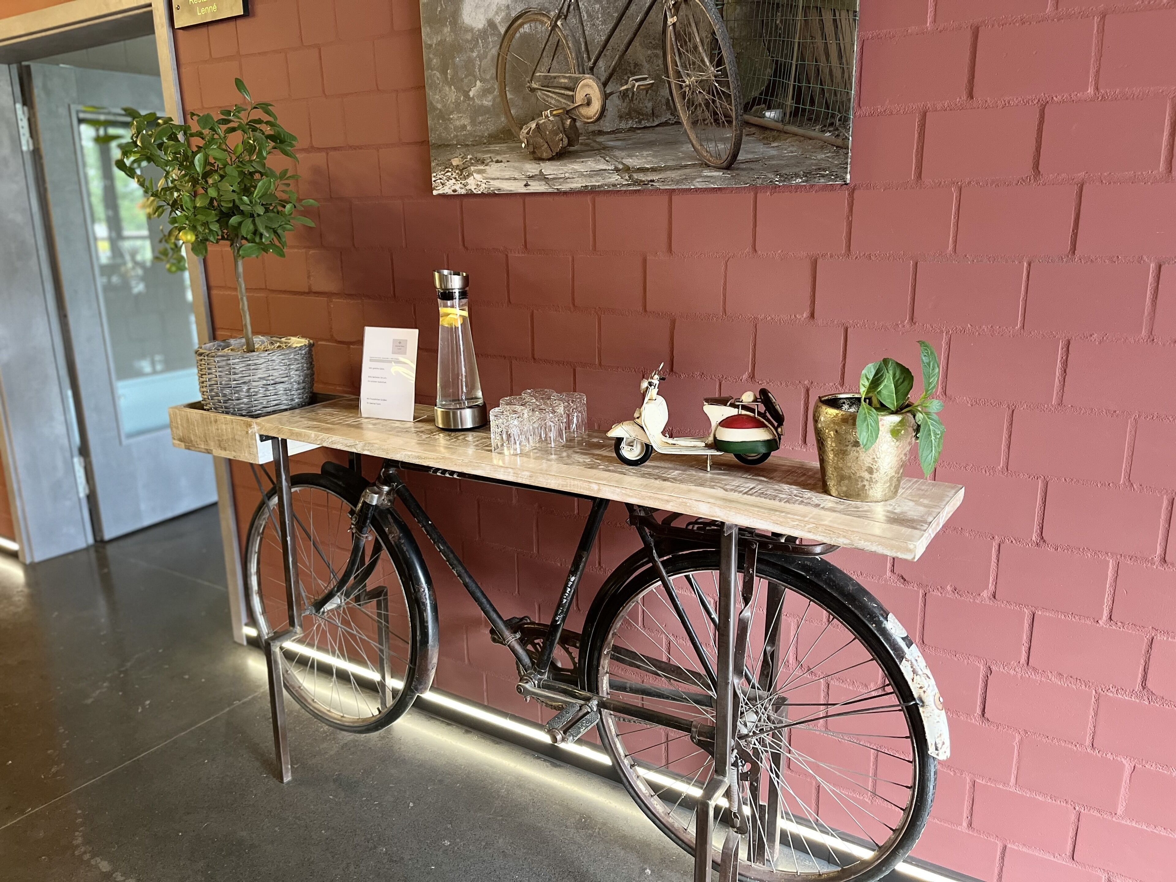 A bicycle serving as a table with a plant, a water bottle, a sign, and a scooter.
