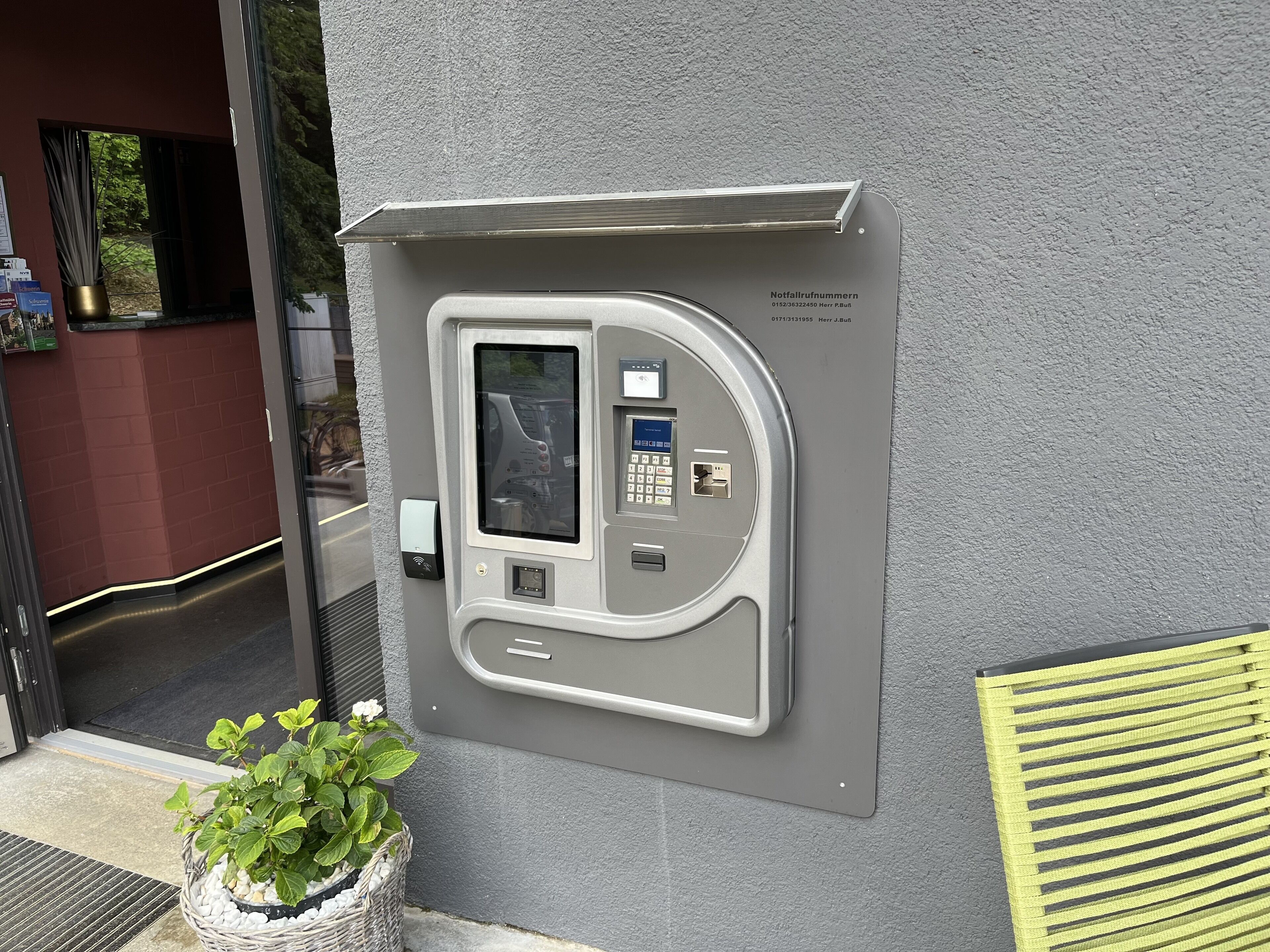 A grey, rectangular, wall-mounted payment kiosk with a screen and keypad, next to a door with a glass panel.