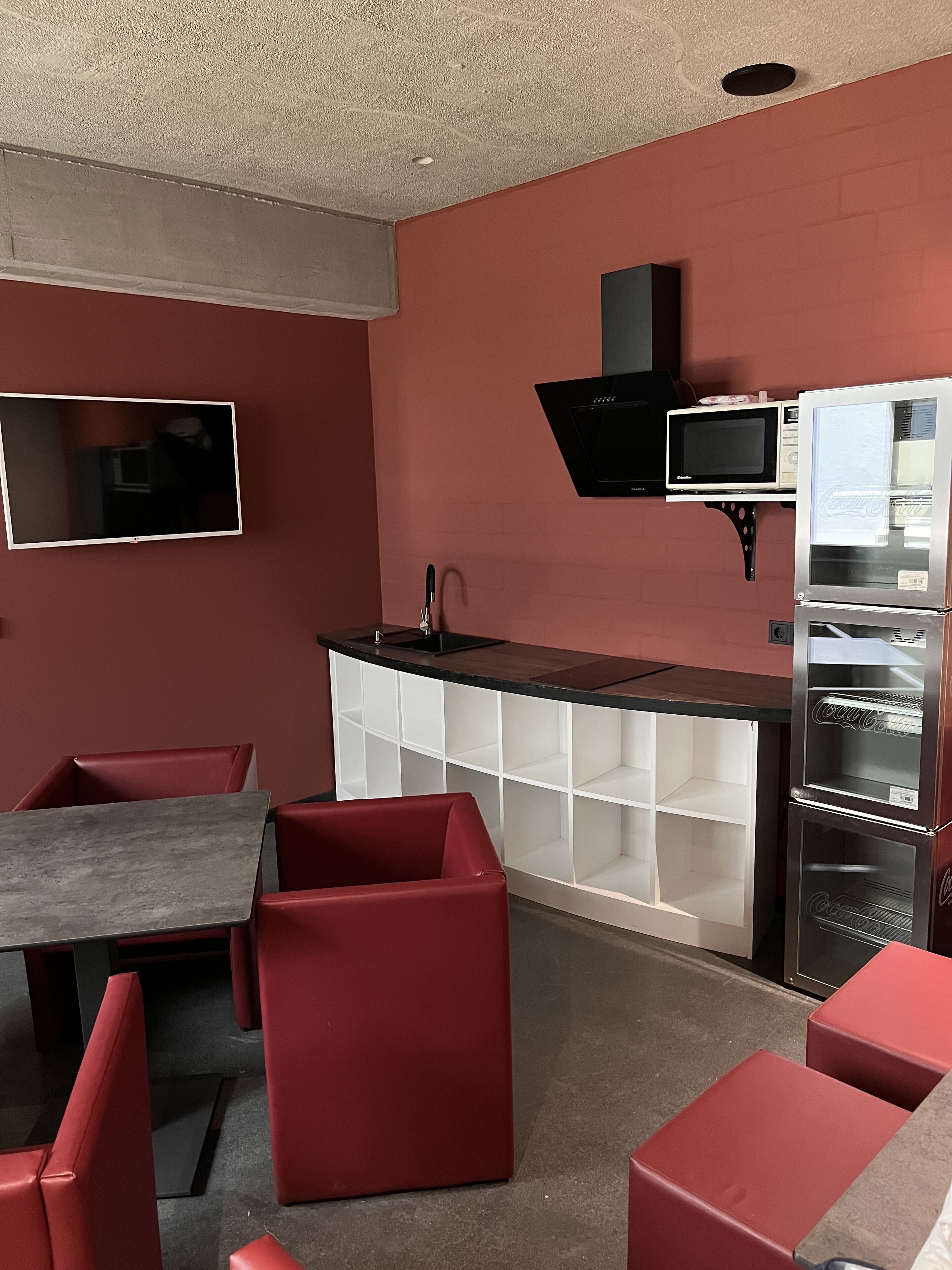 A modern kitchen with a black microwave, a built-in oven, and a refrigerator. There is a bar counter with a sink and a television mounted on the wall. Red chairs and a table are arranged in the dining area.