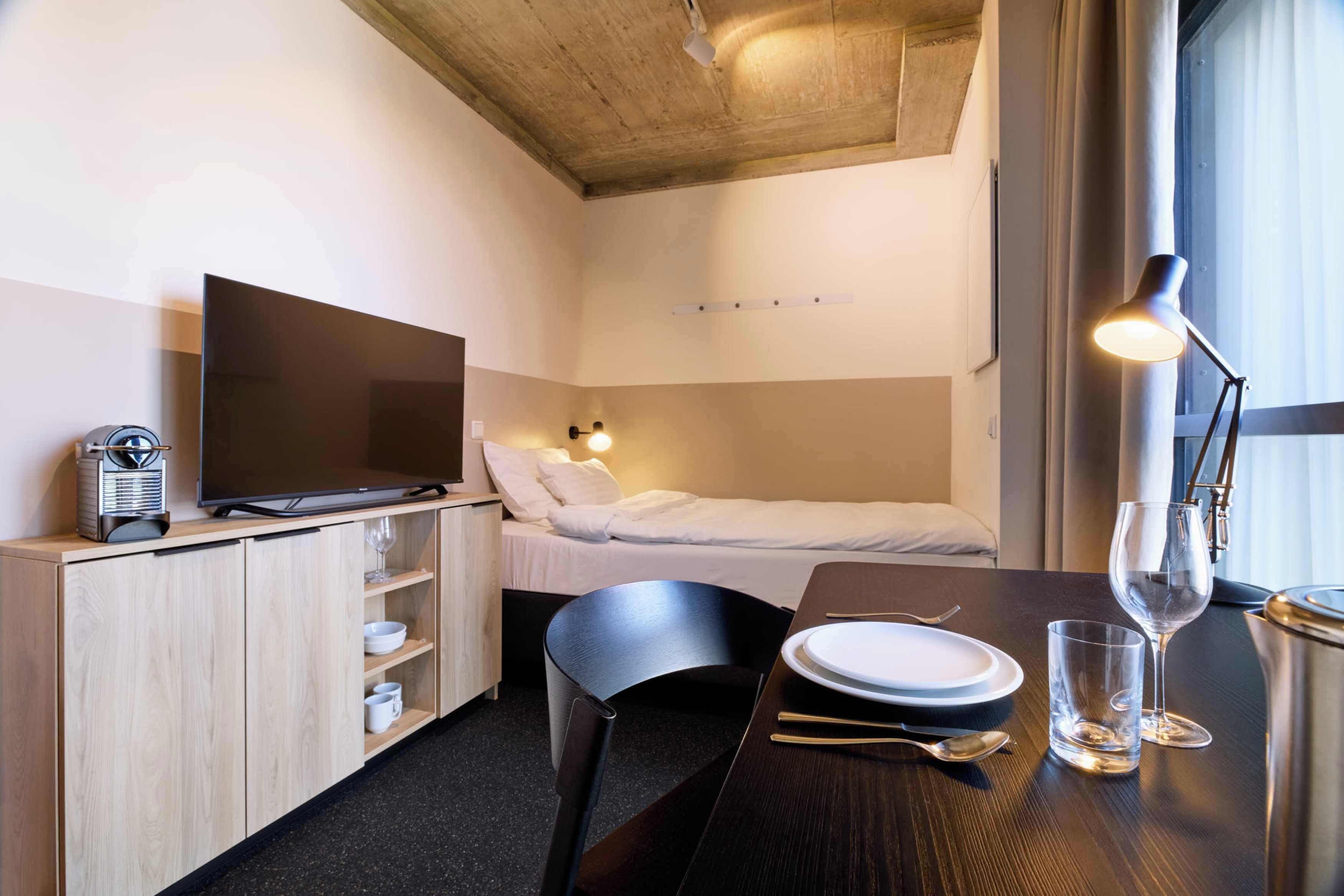 Ein gemütliches Hotelzimmer mit Bett, Schreibtisch mit Stuhl, Fernseher, Nachttisch mit Lampe und einem Fenster mit Vorhängen.