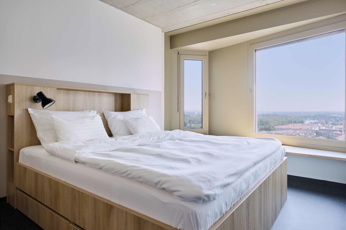 A modern bedroom with a large bed, built-in wooden headboard, a bedside lamp, and a large window offering a city view.