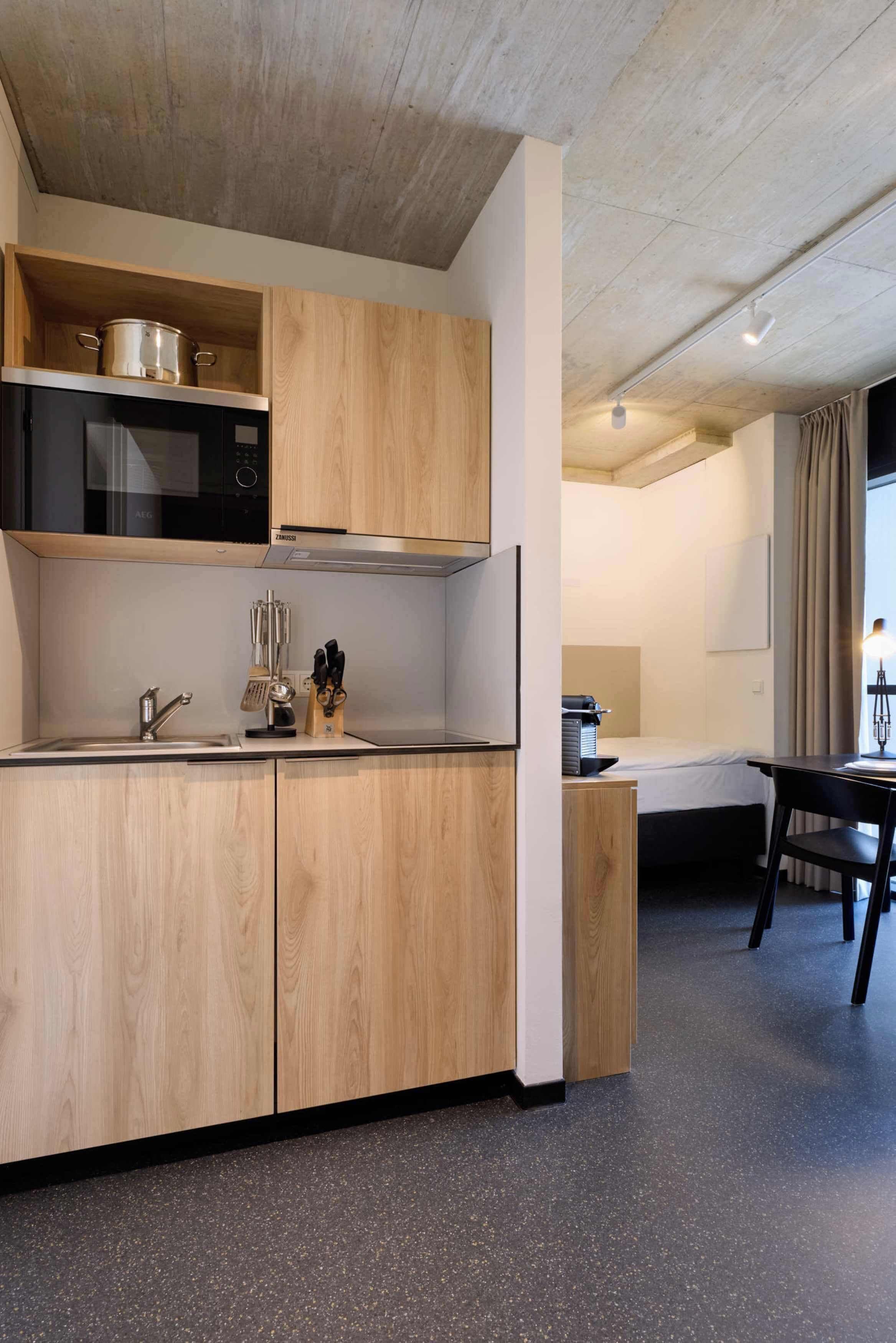 A modern kitchen with wooden cabinets, a stainless steel sink, and a microwave. There is a dining area with a table and chairs, and a window with curtains.