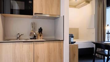 A modern kitchen with wooden cabinets, a stainless steel sink, and a microwave. There is a dining area with a table and chairs, and a window with curtains.