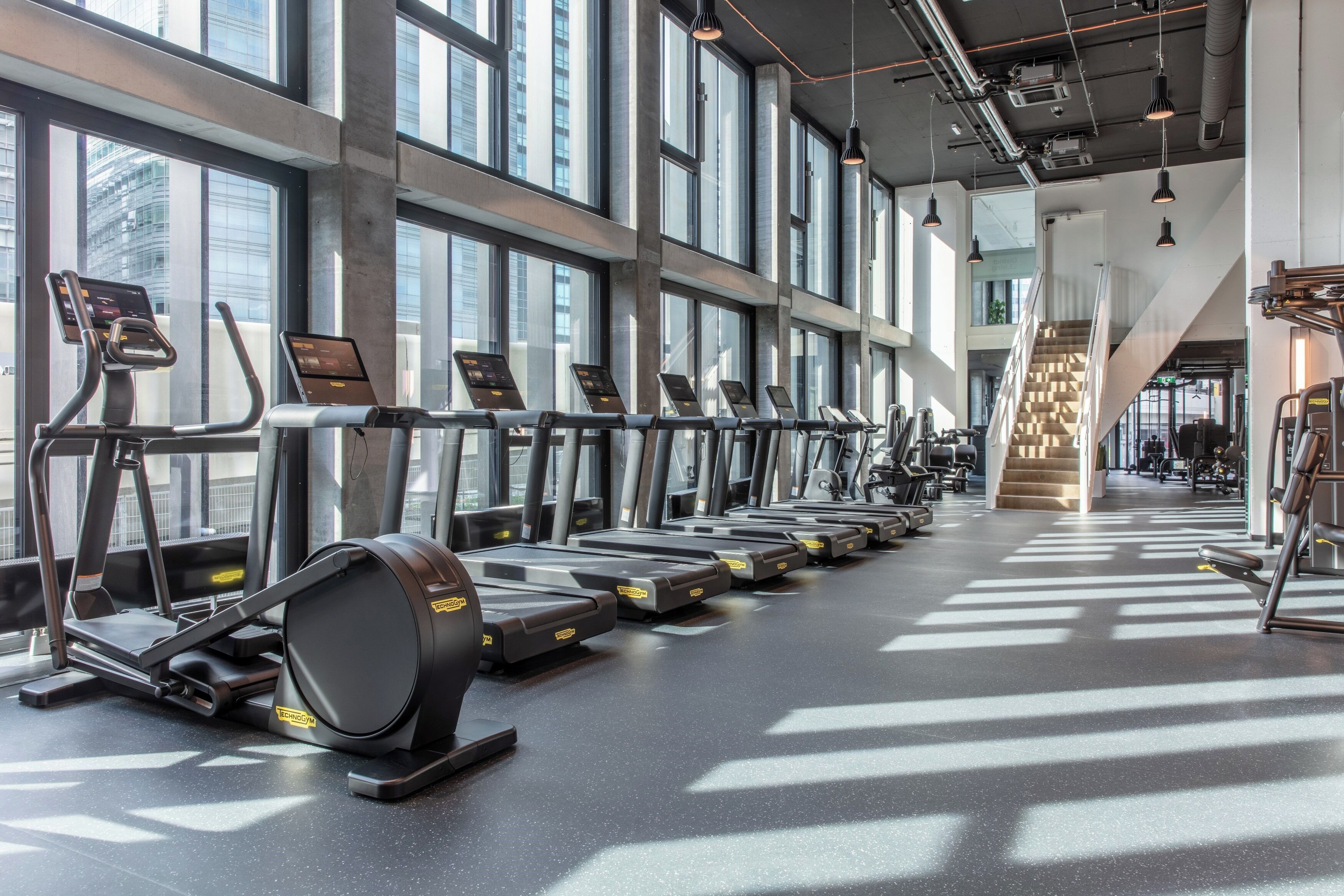 A gym with treadmills aligned along a wall with large windows, a staircase, and various exercise equipment.
