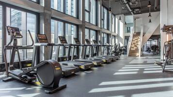 A gym with treadmills aligned along a wall with large windows, a staircase, and various exercise equipment.