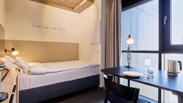 A hotel room with a bed, a desk with a lamp, a chair, a window with curtains, and a view outside.