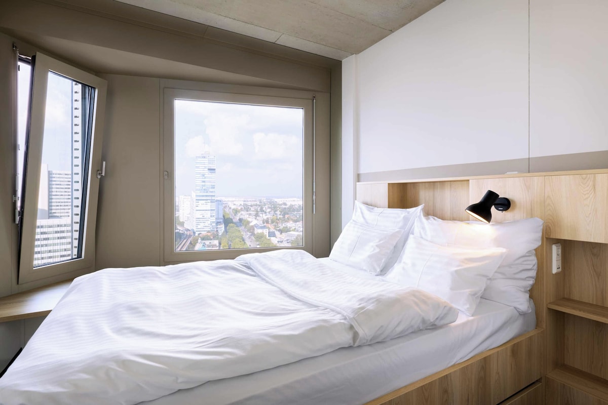 A modern bedroom with a bed, a nightstand, a lamp, and a view of the city through a large window.