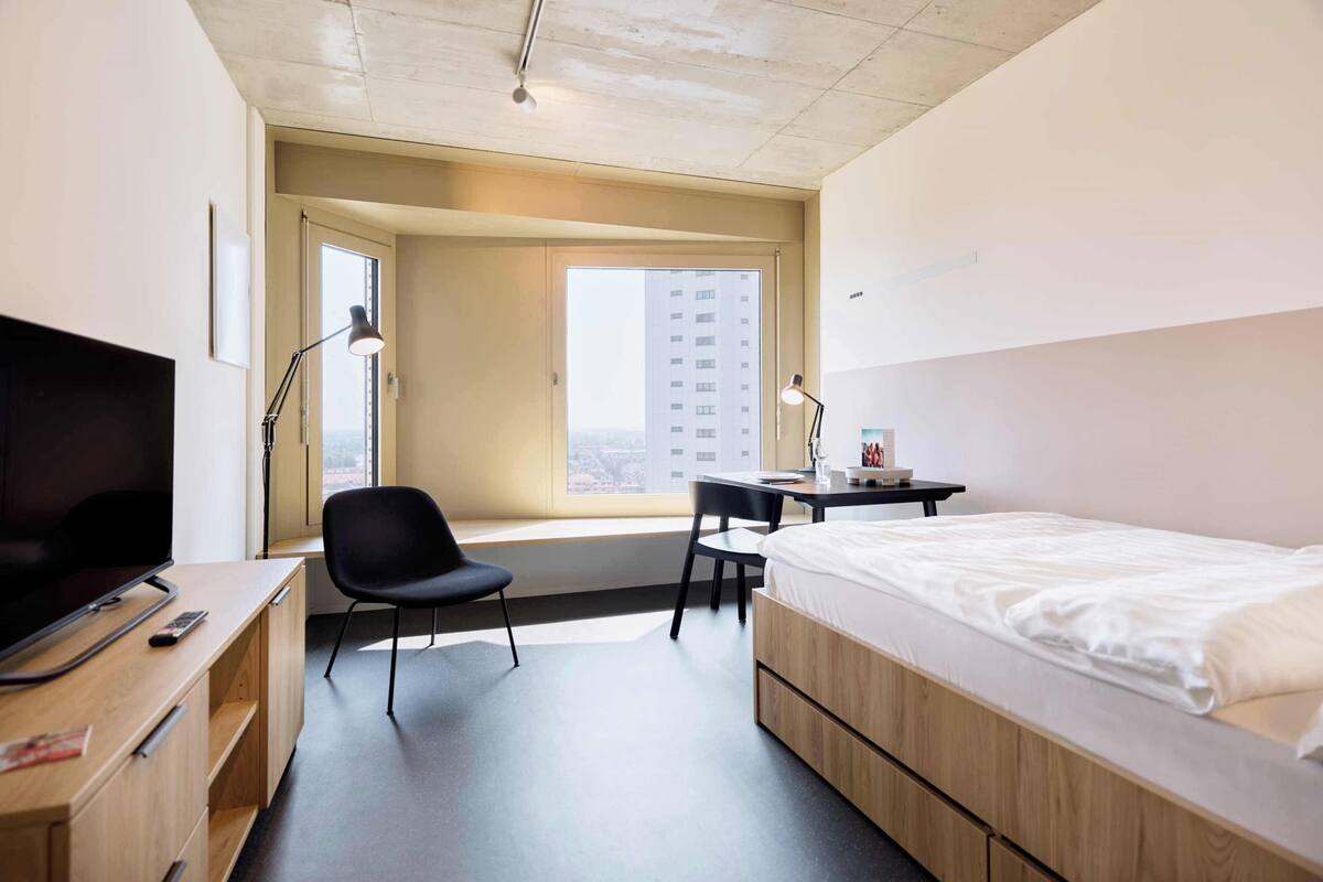 A hotel room with a bed, desk, chair, TV, and a window with a city view.