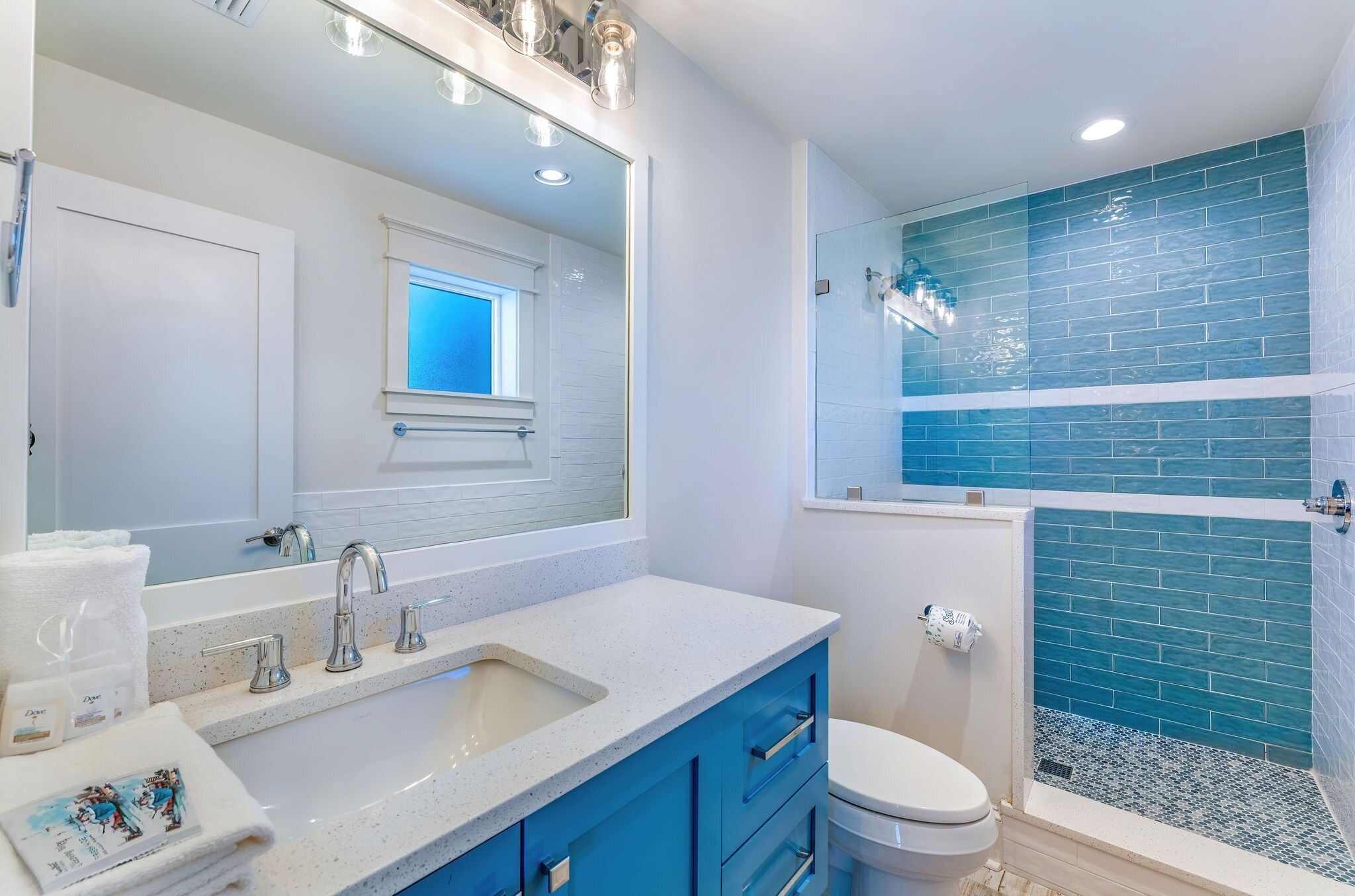 Une salle de bain avec un lavabo blanc, des meubles bleus, des toilettes blanches et une douche en verre, dont les parois sont recouvertes de carreaux bleus.