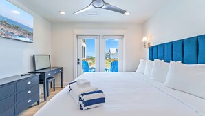 Une chambre d’hôtel équipée d’un lit, d’un bureau, d’une chaise, d’un ventilateur de plafond et offrant une vue sur la plage et les bateaux par les fenêtres.