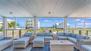 Un espace extérieur aménagé, abrité et doté de meubles en osier et d’un tapis à motifs, offrant une vue sur une plage et des bâtiments.