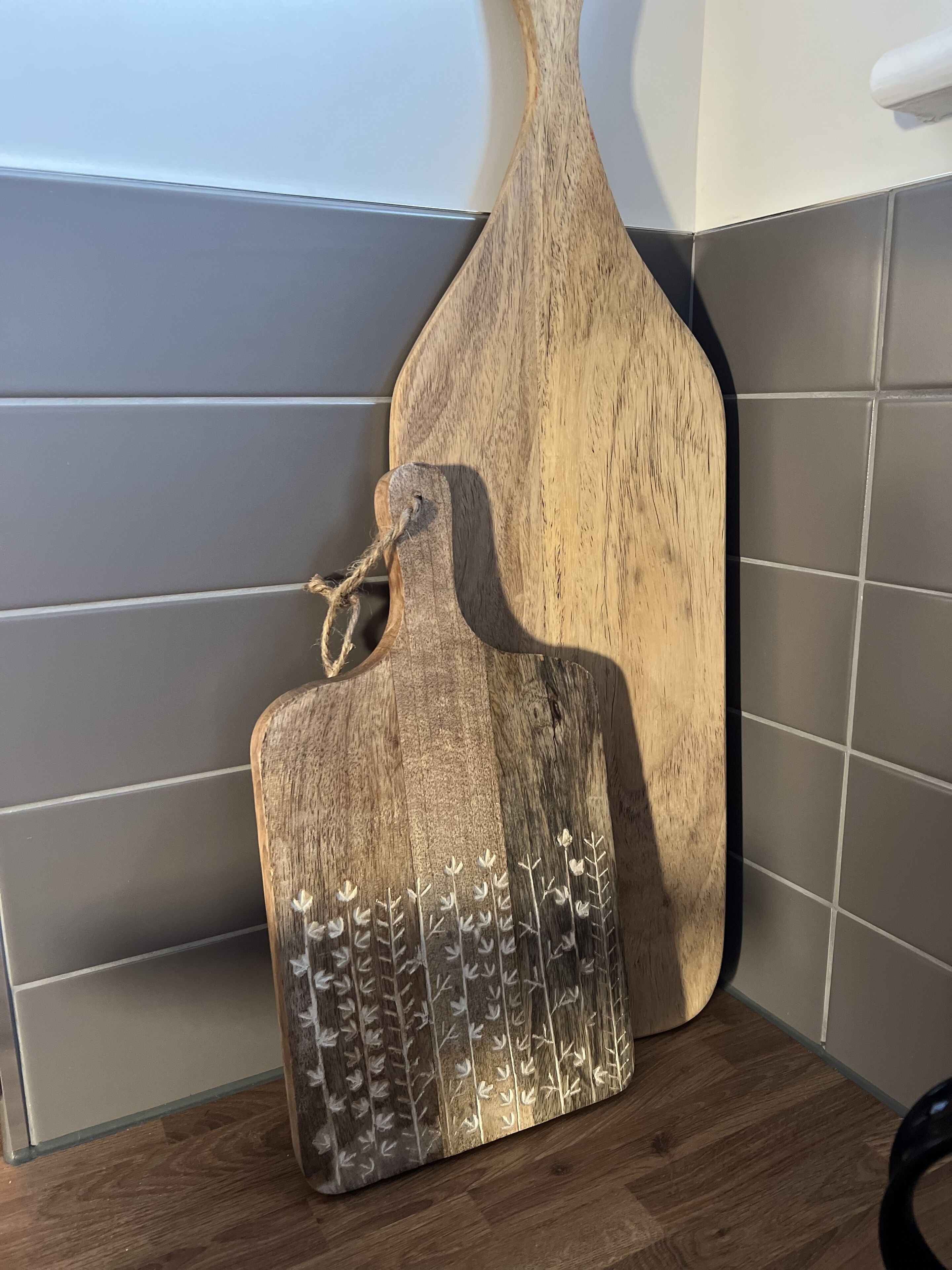 A wooden cutting board with a handle and a decorative pattern, placed on a tiled floor next to a wall.