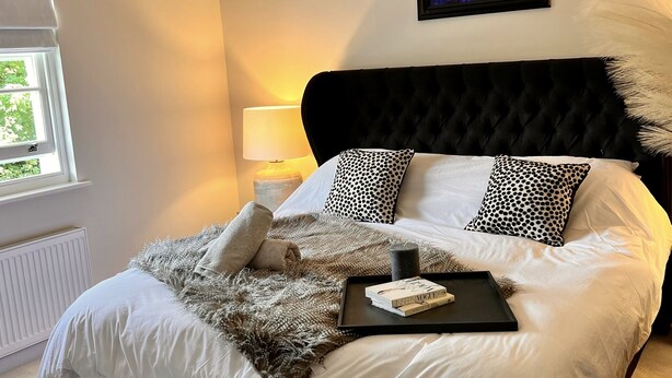 Una cama bien tendida con cabecera negra, ropa de cama blanca y almohadas estampadas. Una bandeja con libros está colocada sobre la cama.