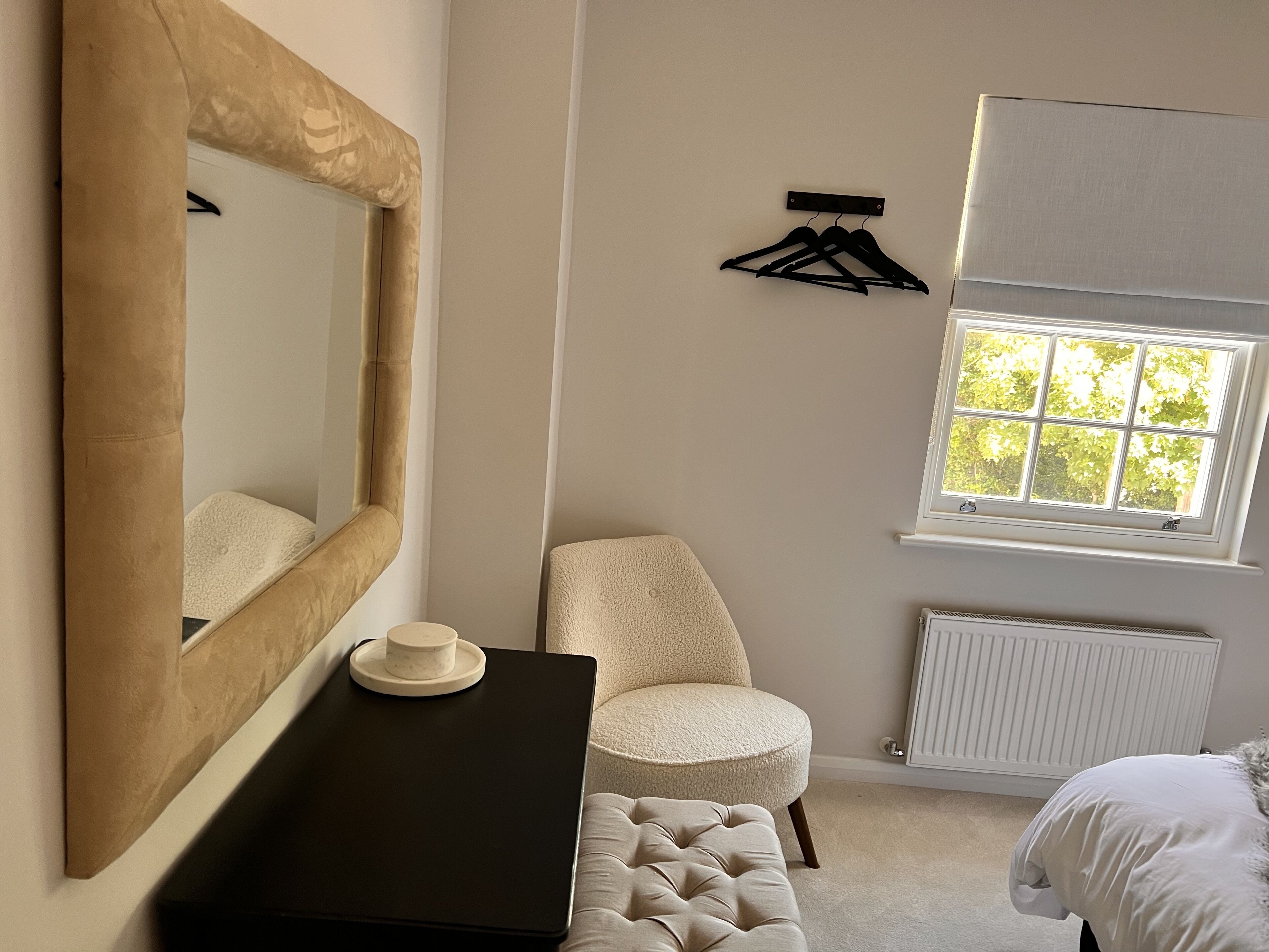 A bedroom with a large mirror, a round chair, a bedside table, a bed, a window with a view of trees, and a radiator.
