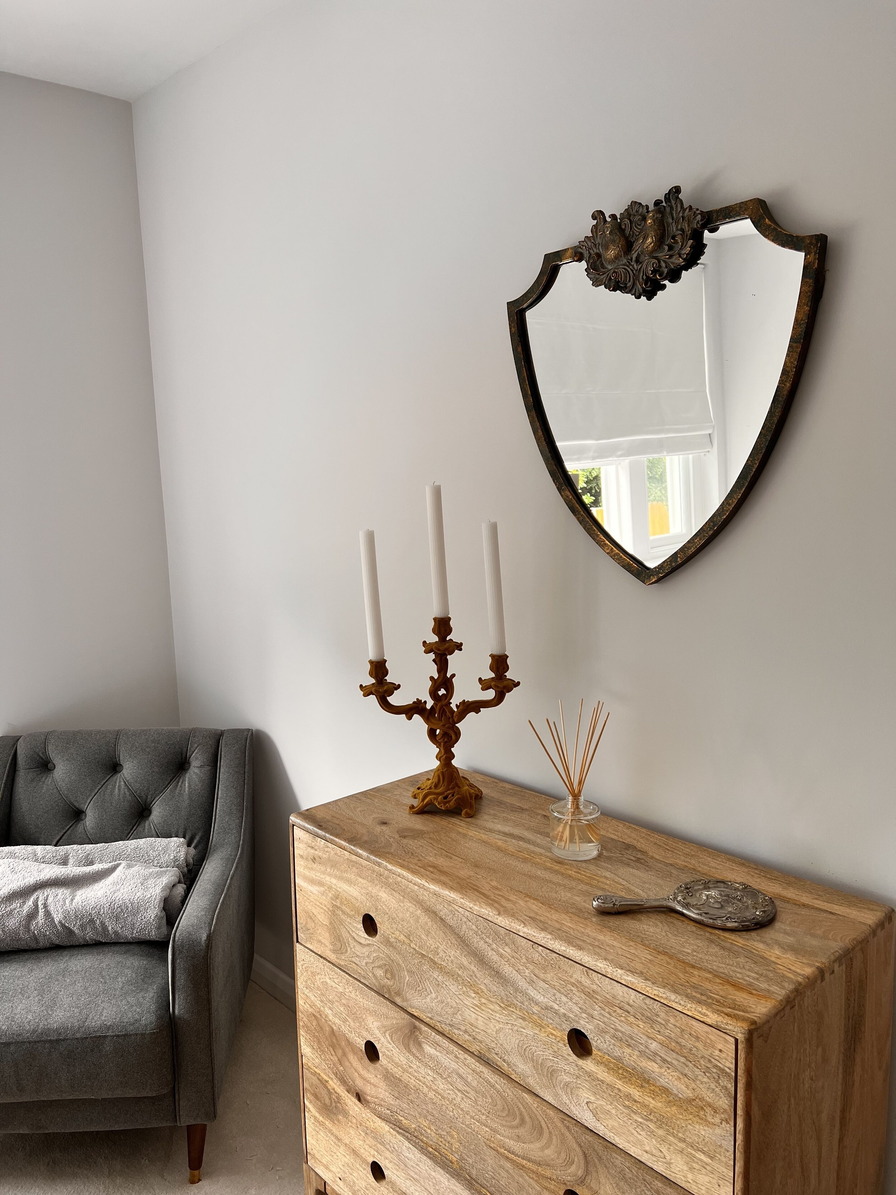 Ein Zimmer mit grauem Sofa, einem Holzkleiderschrank, einem Kerzenleuchter mit Kerzen, einem Spiegel und Reisediffusor-Stäbchen.