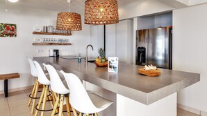 Cozinha moderna com ilha central, banquetas brancas, geladeira de aço inoxidável e luminárias de vime decorativas.