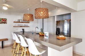 Eine moderne Küche mit einer zentralen Kochinsel, weißen Barhocker, einem Edelstahlkühlschrank und dekorativen Korbtassenlampen.