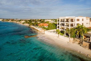 Ein Küstenhotel mit Blick auf den Strand, das ein mehrstöckiges Gebäude mit Balkonen, einen Sandstrand und klares, türkisblaues Wasser zeigt.