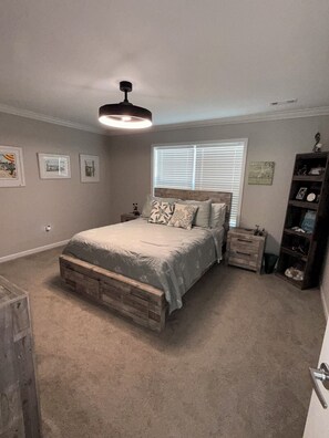 Une chambre à coucher comprenant un lit, des tables de chevet, une commode, un banc en bois, un luminaire au plafond, une fenêtre avec des stores et des éléments de décoration murale.