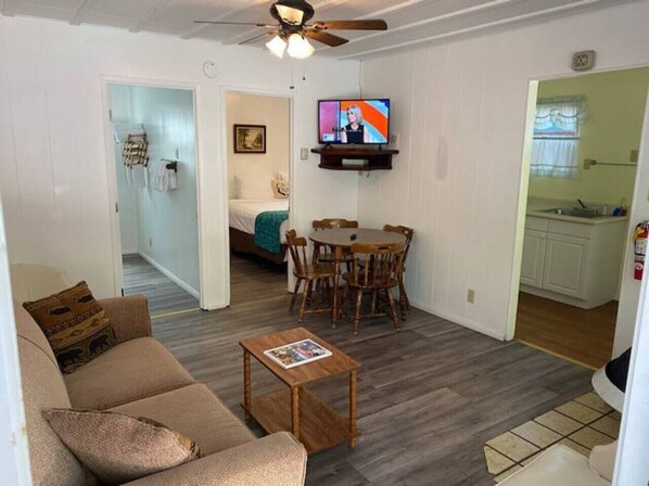 Sala de estar con sofá, mesa de centro, televisor, mesa de comedor y sillas. Se ve una cocina a través de un acceso abierto.