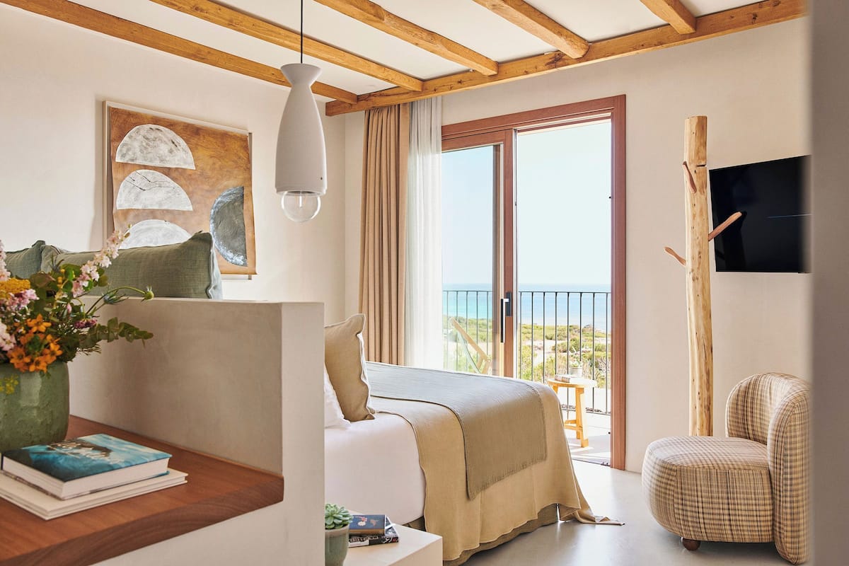 A bedroom with a bed, a chair, a table with books and a vase of flowers, a painting, and a balcony with a view.