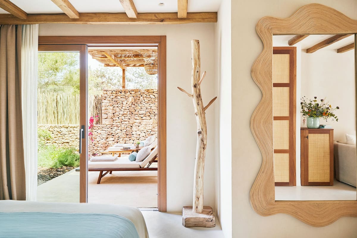 A bedroom with a bed, a wooden beam ceiling, a stone wall, and a view of an outdoor seating area.