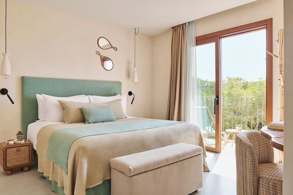 A bedroom with a large bed, a bench, a bedside table, a mirror, and a view of the outdoors.