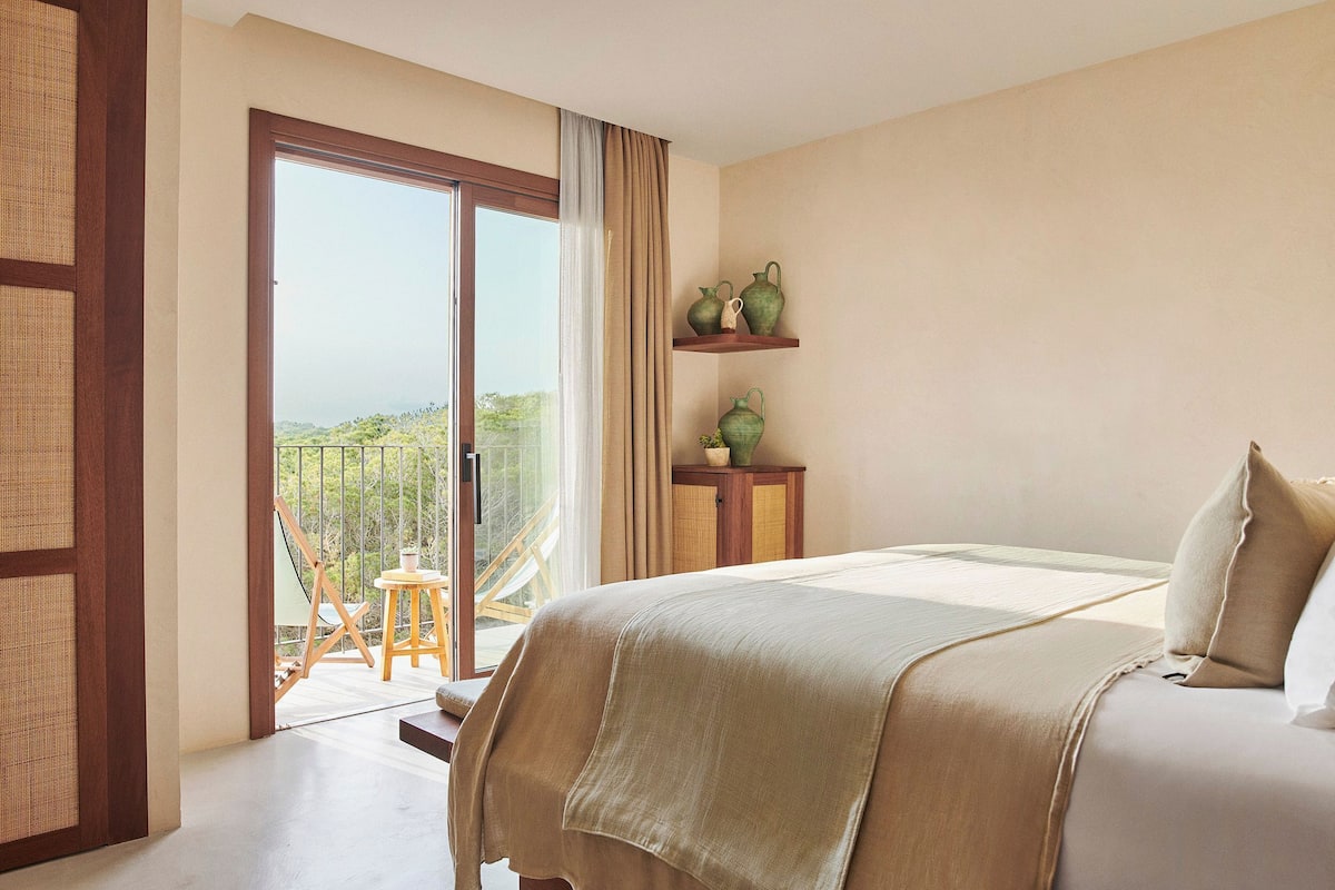 A bedroom with a large bed, a balcony with a view, and a small table with decorative items.