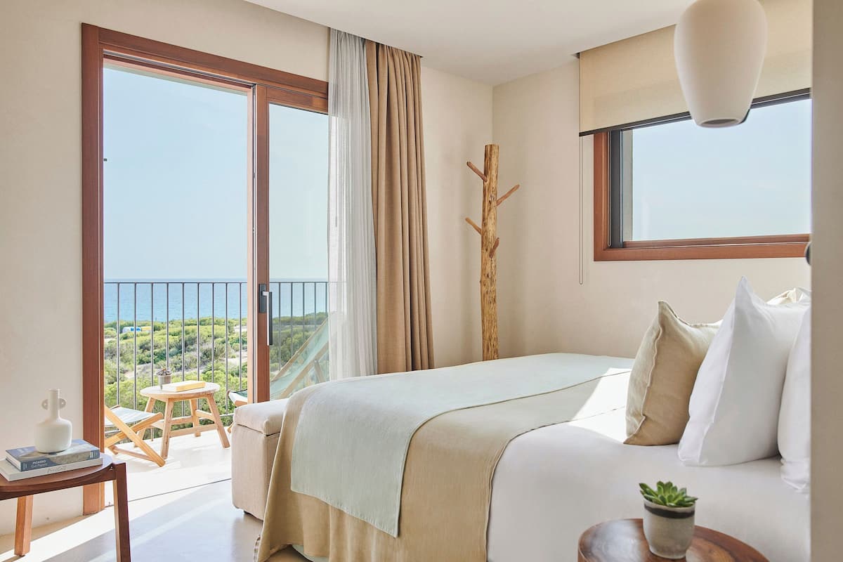 A bedroom with a bed, a small table, a chair, a potted plant, and a view of the outdoors.