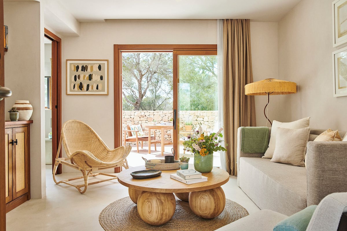 A living room with a wicker chair, a round wooden coffee table, a sofa, and a view through a glass door to an outdoor area.