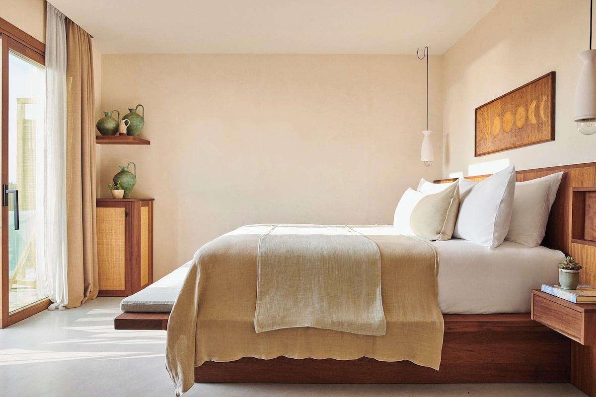 A neatly arranged bedroom with a bed, bedside tables, a wall-mounted shelf with decorative items, and a large window with curtains.