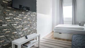 A modern bedroom with a stone wall, a mounted TV, a bed with white bedding, and a grey upholstered bench. - Großzügige Wohnung in Berlin Mitte für 6 Gäste (Berlin)