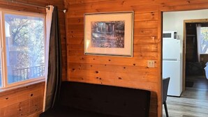 Interior de una cabaña de madera con un sofá negro, un cuadro en la pared y una ventana con cortinas.