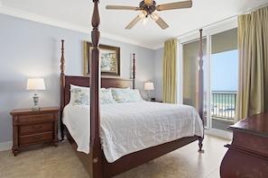 Un letto a baldacchino con ventilatore a soffitto, due lampade da comodino, un comodino e vista sulla spiaggia tramite una porta scorrevole in vetro.