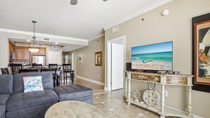 A living room with a gray sofa, a round coffee table, a television on a white console, and a kitchen in the background. - Azure 414 - 3 Bd Beachfront w/ Premium Amenities - Sleeps 8! (Fort Walton Beach)