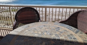 A balcony with wicker chairs and a stone-tiled table overlooking a beach.